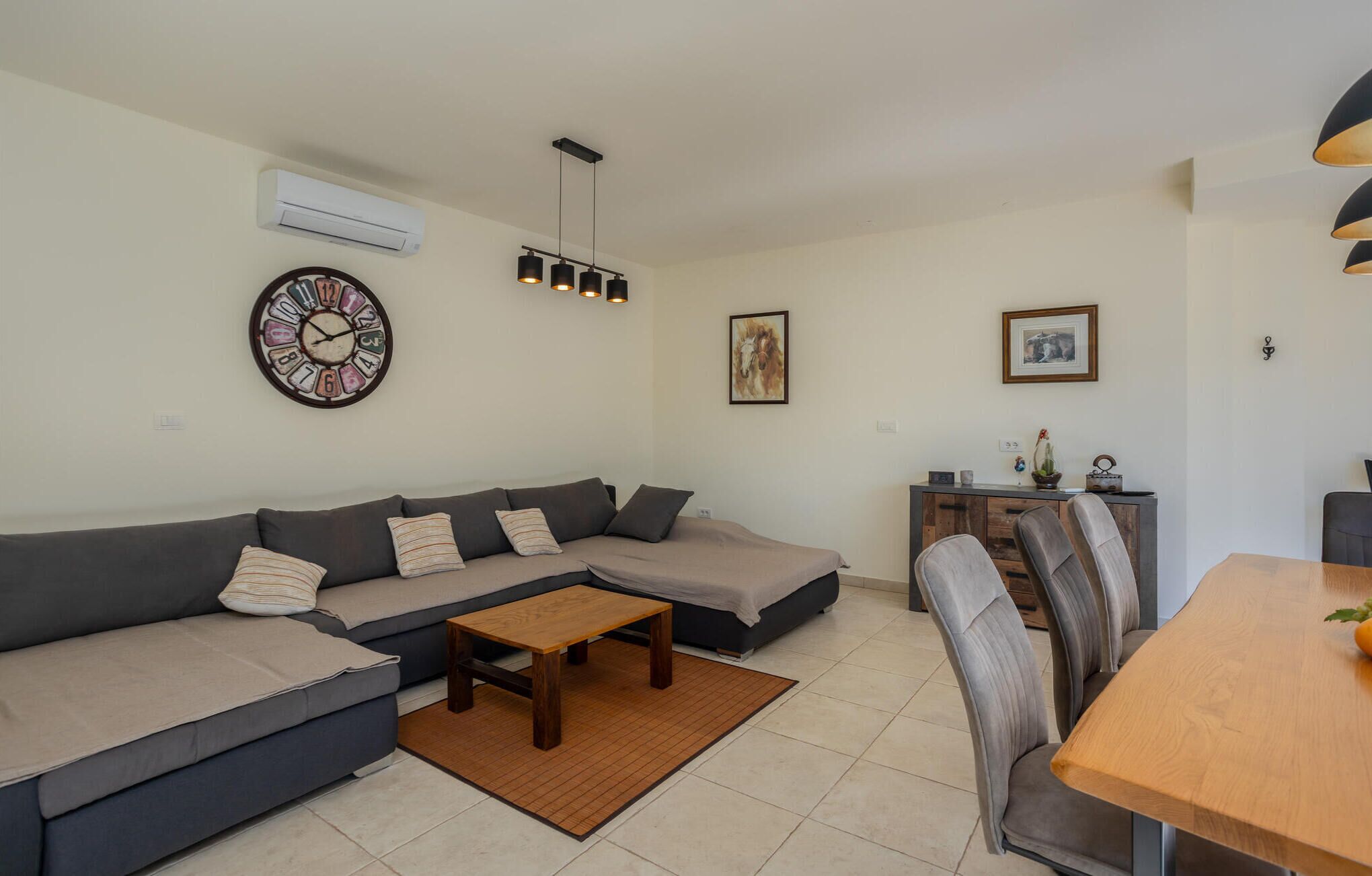 Ein modernes Wohnzimmer mit grauem Sofa, einem Holztisch und einem Essbereich mit Holztisch und grauen Stühlen. An der Wand hängt eine Uhr, und ein kleines Sideboard ist mit Dekorationen geschmückt.