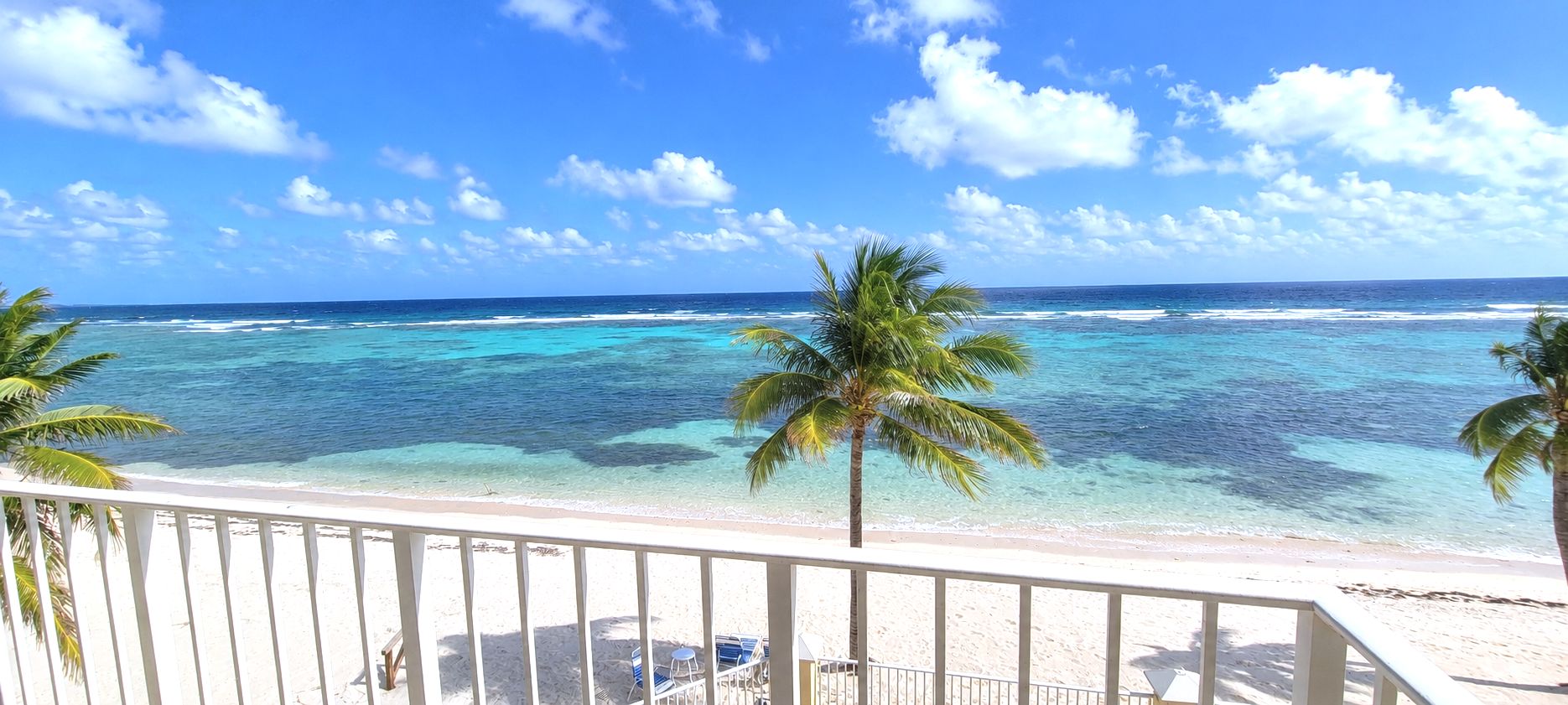 A beach view with clear turquoise water, palm trees, and a white balcony railing.