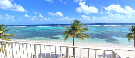 A beach view with clear turquoise water, palm trees, and a white balcony railing.