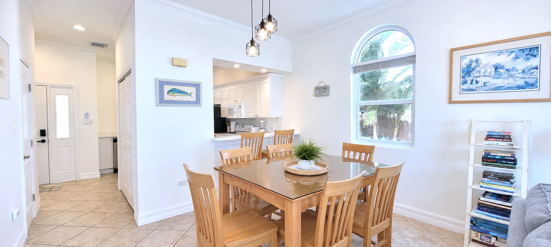 A dining area with a glass-top table and wooden chairs, a kitchen with white cabinets, a bookshelf, and a large window with a view of palm trees.
