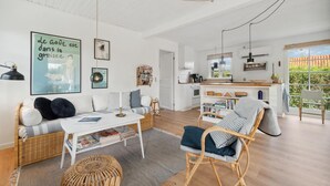 Un salon spacieux avec un canapé, une table basse et des fauteuils en osier. On aperçoit une cuisine à l’arrière-plan, avec un coin repas.