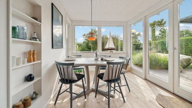 Un coin repas avec une table et des chaises blanches, de grandes fenêtres et une vue sur la verdure et un voilier à l’extérieur.