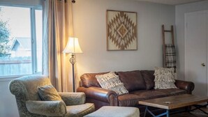 Sala de estar com sofá de couro marrom, poltrona estampada, mesa de centro de madeira, luminária de chão e uma escada encostada na parede.