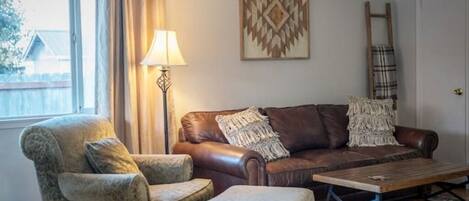 A living room with a brown leather sofa, a patterned armchair, a wooden coffee table, a floor lamp, and a ladder leaning against the wall.