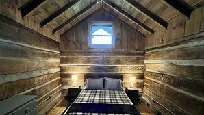 Un dormitorio rústico de cabaña de madera con una cama, dos mesitas de noche con lámparas, una ventana y un aparador de madera.