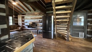 Interior rústico de una cabaña con techo de madera, chimenea de piedra, área de cocina con estufa, refrigerador y escalera de madera.