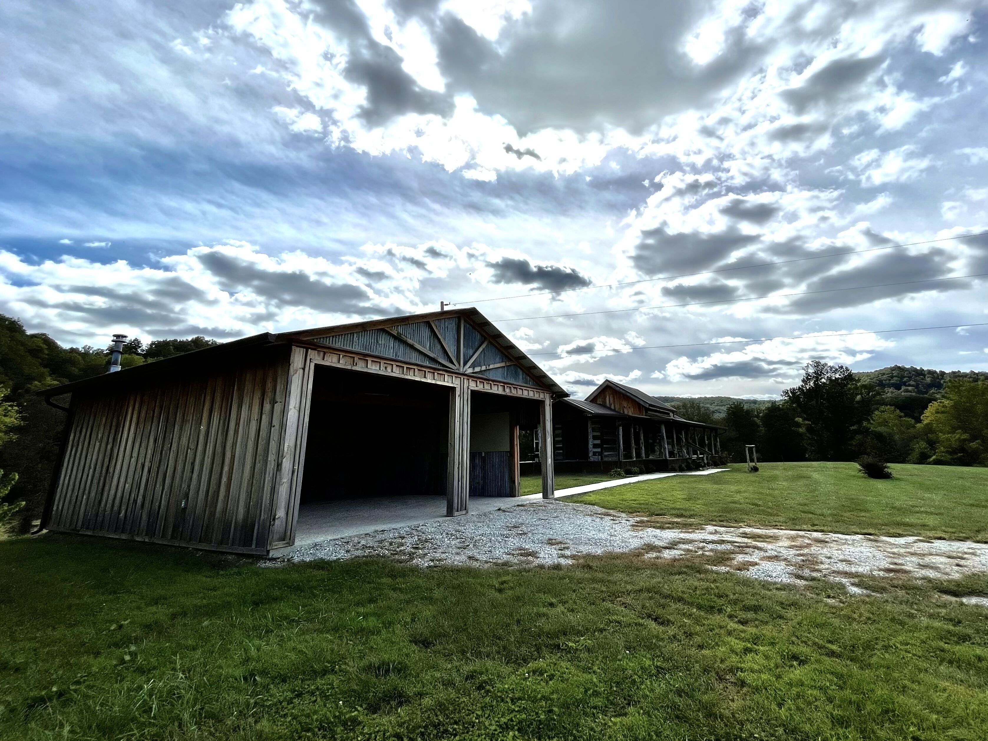 Un bâtiment de style rustique, évoquant une grange, doté d’une grande porte de garage ouverte, et entouré d’une zone herbeuse et d’arbres.