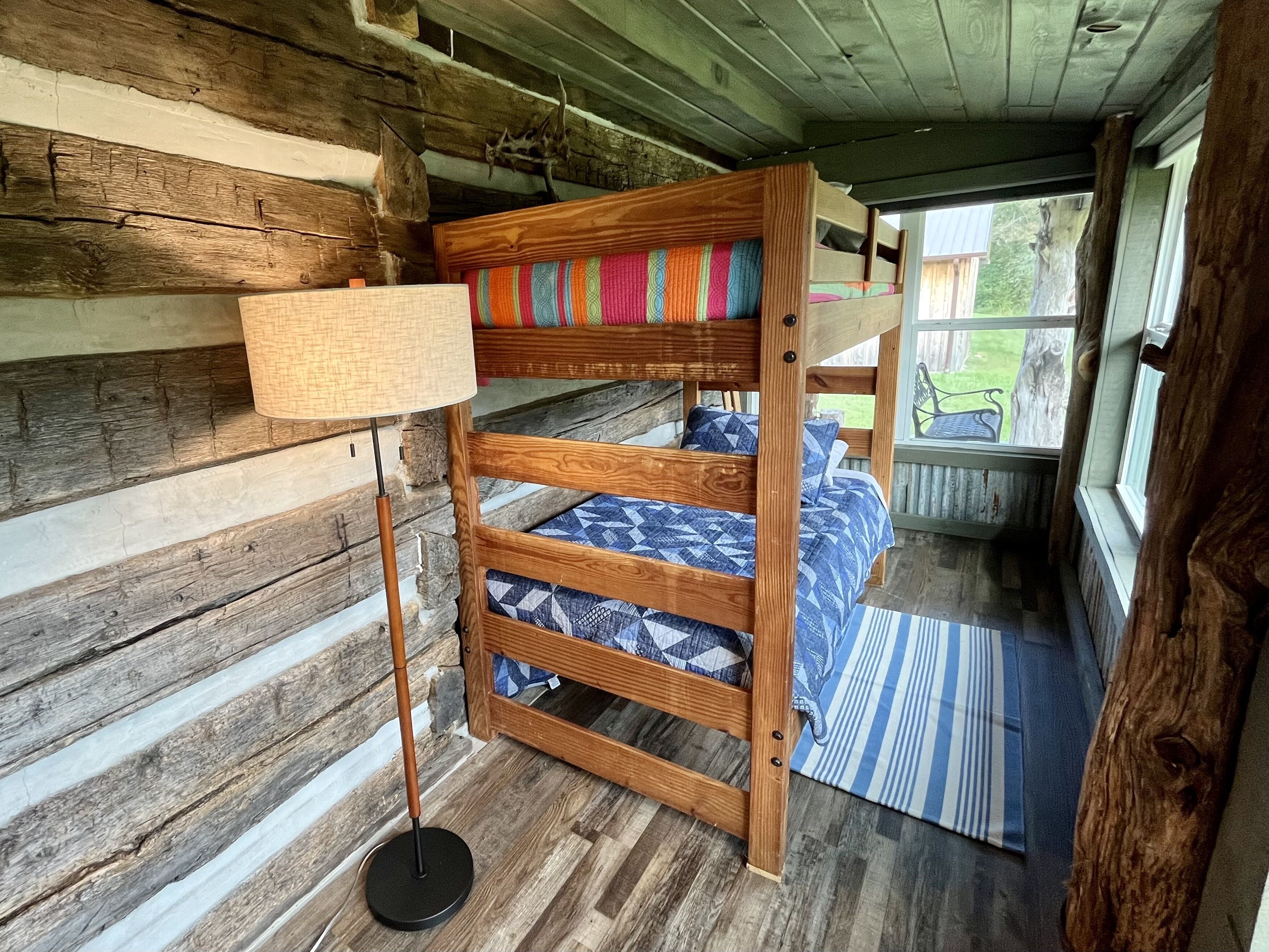 A bunk bed with colorful bedding, a floor lamp, and a view of the outdoors through a window.