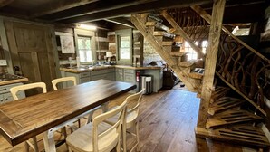 Una cocina rústica con pisos de madera, una mesa de comedor con sillas, una escalera con baranda de madera y vistas al exterior a través de una ventana.