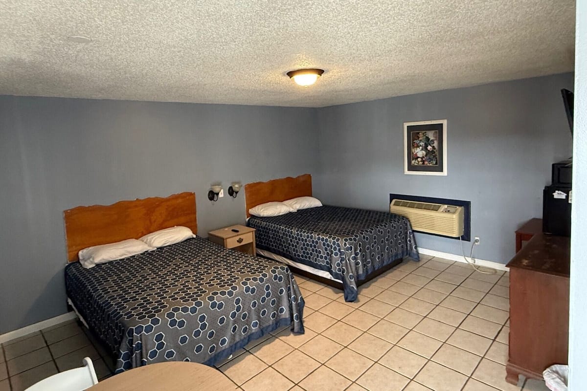 A hotel room with two beds, a small table, a chair, a wall-mounted air conditioner, and a ceiling light.