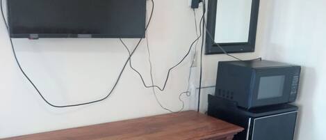 A room with a wooden dresser, a flat-screen TV mounted on the wall, a small black refrigerator, and a wall-mounted mirror.