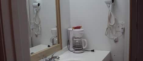 Un baño con un lavamanos, dos secadores de pelo fijados en la pared y una cafetera sobre la encimera.