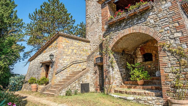 Un bâtiment rustique en pierre, doté d’un petit escalier menant à une porte en bois, le tout entouré de verdure et de plantes en pot.