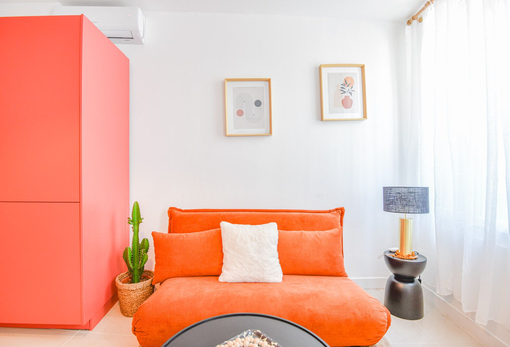 Une pièce avec un canapé rouge, un coussin blanc, un tableau encadré au mur, un cactus et une lampe sur une table d’appoint.