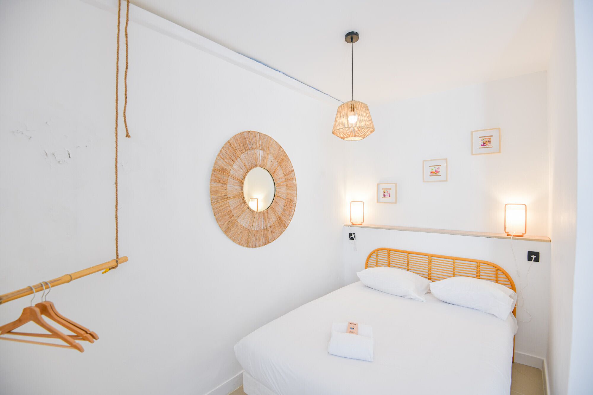 A bedroom with a bed, a hanging light fixture, a wall-mounted mirror, and a bedside table with a lamp.