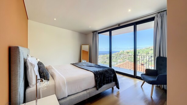A modern bedroom with a large bed, a bedside table, a chair, and a view of the sea through large windows. - Dream House 2, by LSA Madeira (Calheta)
