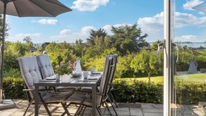 An outdoor dining area with a table and chairs, a large umbrella, and a view of a lush garden and distant landscape.
