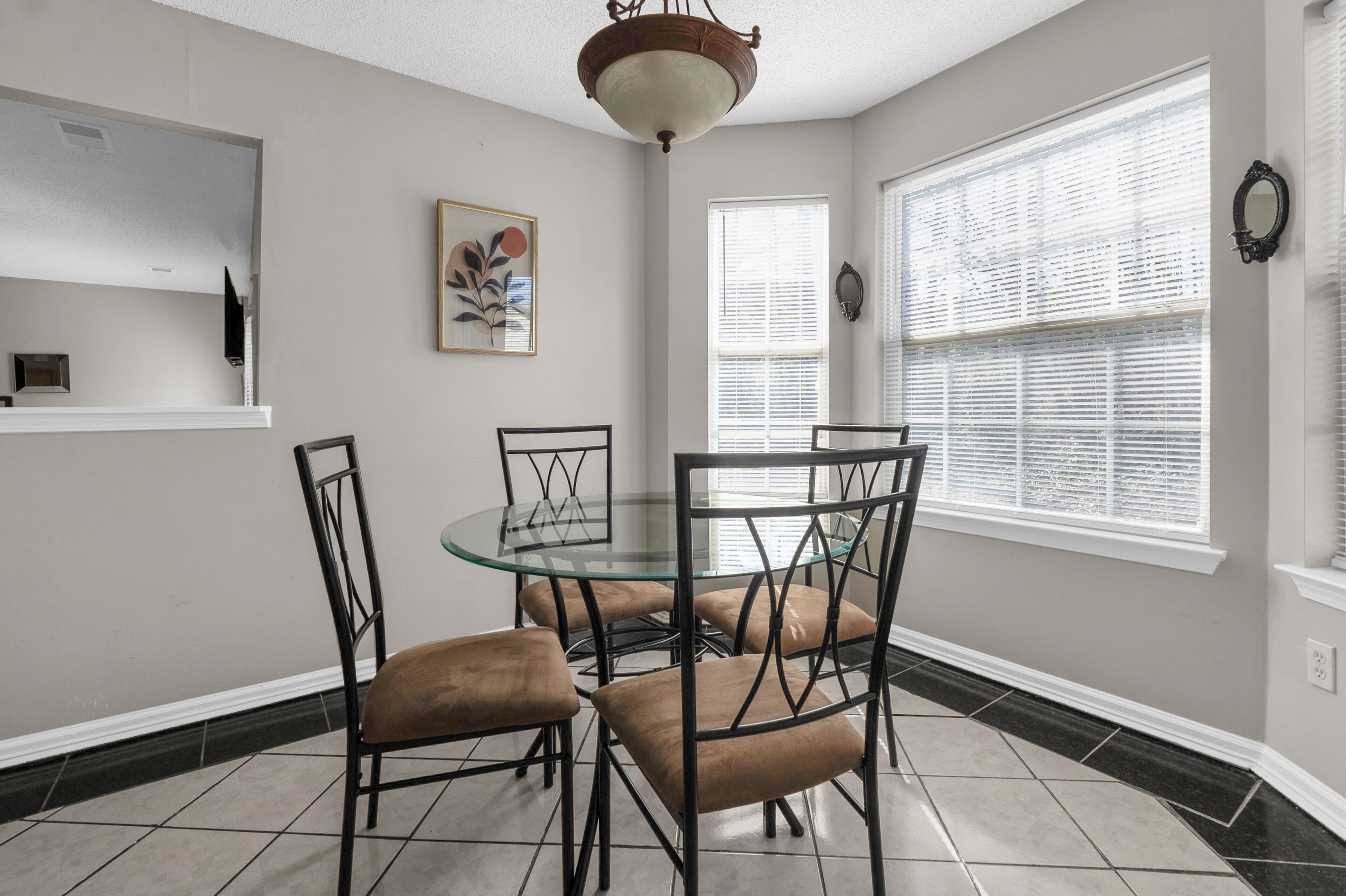 A dining area with a glass table and four chairs, a ceiling light fixture, and a window with blinds.
