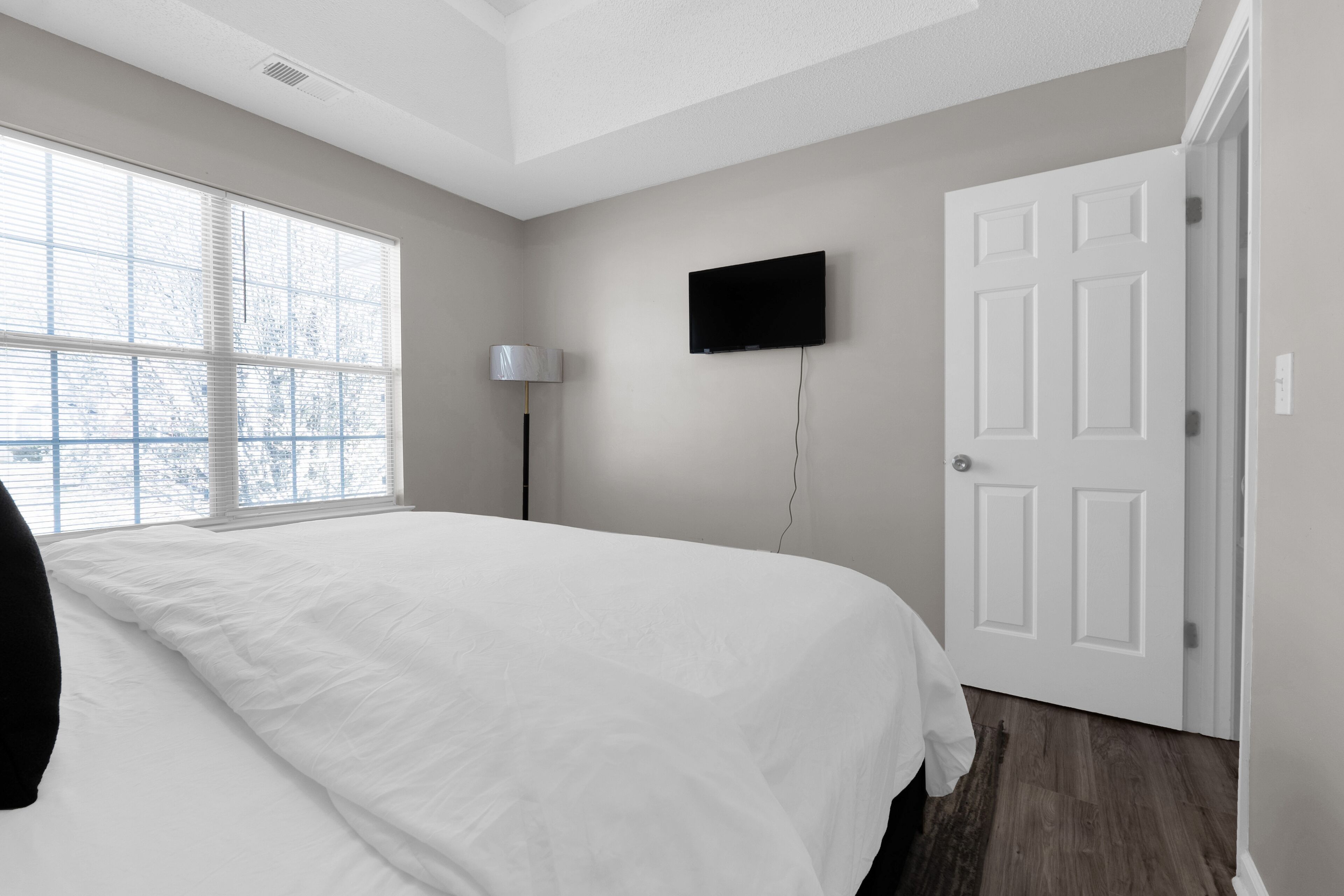 A bedroom with a large bed, a flat-screen TV mounted on the wall, a door, and a window with blinds.
