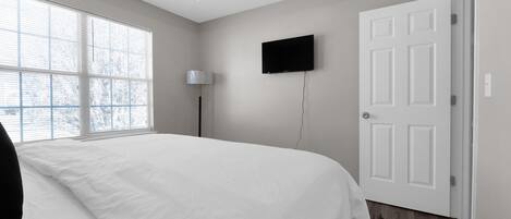 A bedroom with a large bed, a flat-screen TV mounted on the wall, a door, and a window with blinds.