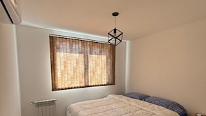 Een slaapkamer met een bed, een radiator, een raam met zonwering, een hanglamp en een nachtkastje.
