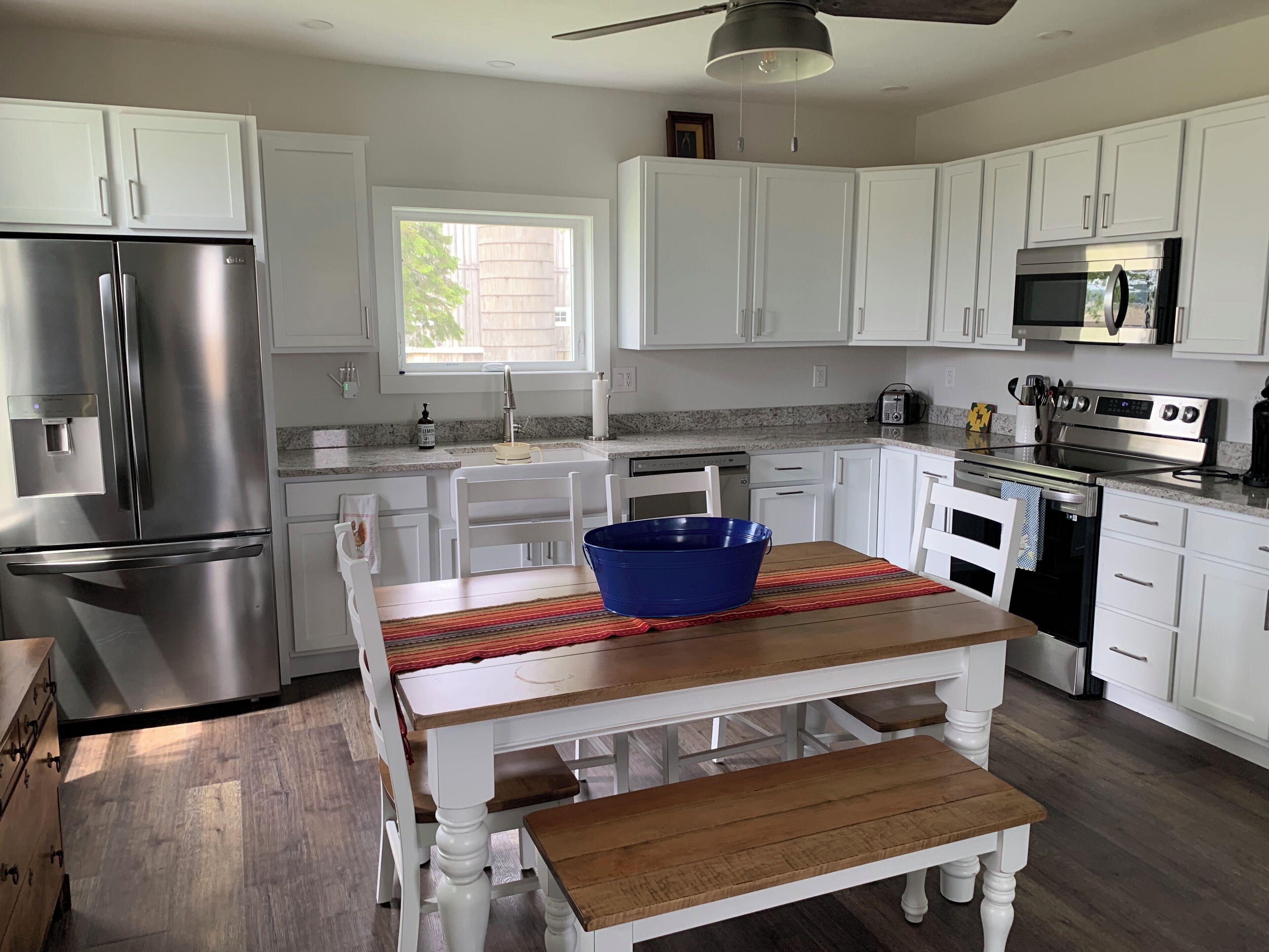 A modern kitchen with white cabinets, stainless steel appliances, a wooden dining table with a blue bowl, and a window providing natural light.