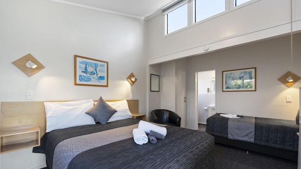 A hotel room with a large bed, a smaller bed, a desk, and a chair. There are two paintings on the walls and a window above the beds. - Central Inn Taupo (Taupo)