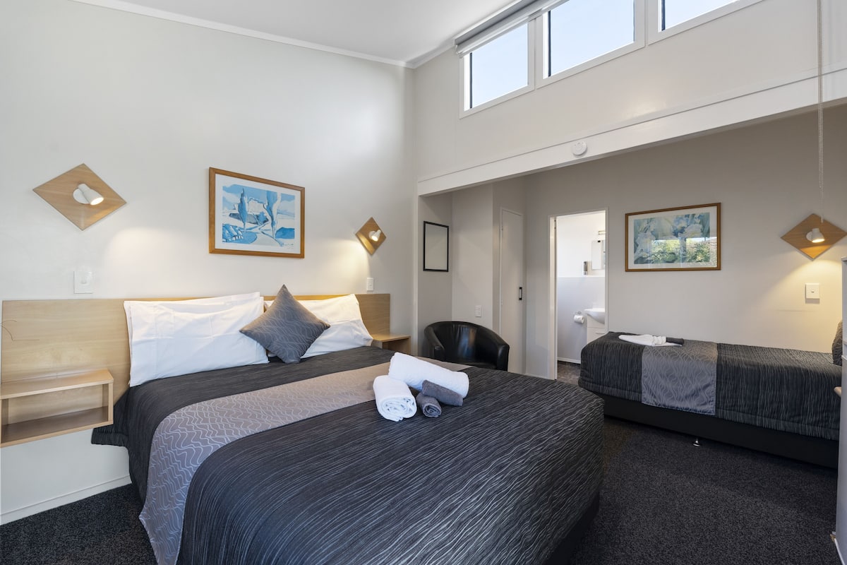 Ein Hotelzimmer mit einem großen Bett, einem kleineren Bett, einem Schreibtisch und einem Stuhl. An den Wänden hängen zwei Gemälde und über den Betten befindet sich ein Fenster.