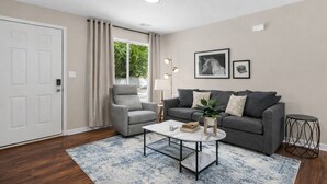 Una sala de estar moderna con un sofá gris, un sillón gris, una mesa de centro blanca y un tapete azul estampado. Hay una ventana con cortinas, un cuadro enmarcado en la pared y una mesita de noche con una lámpara.