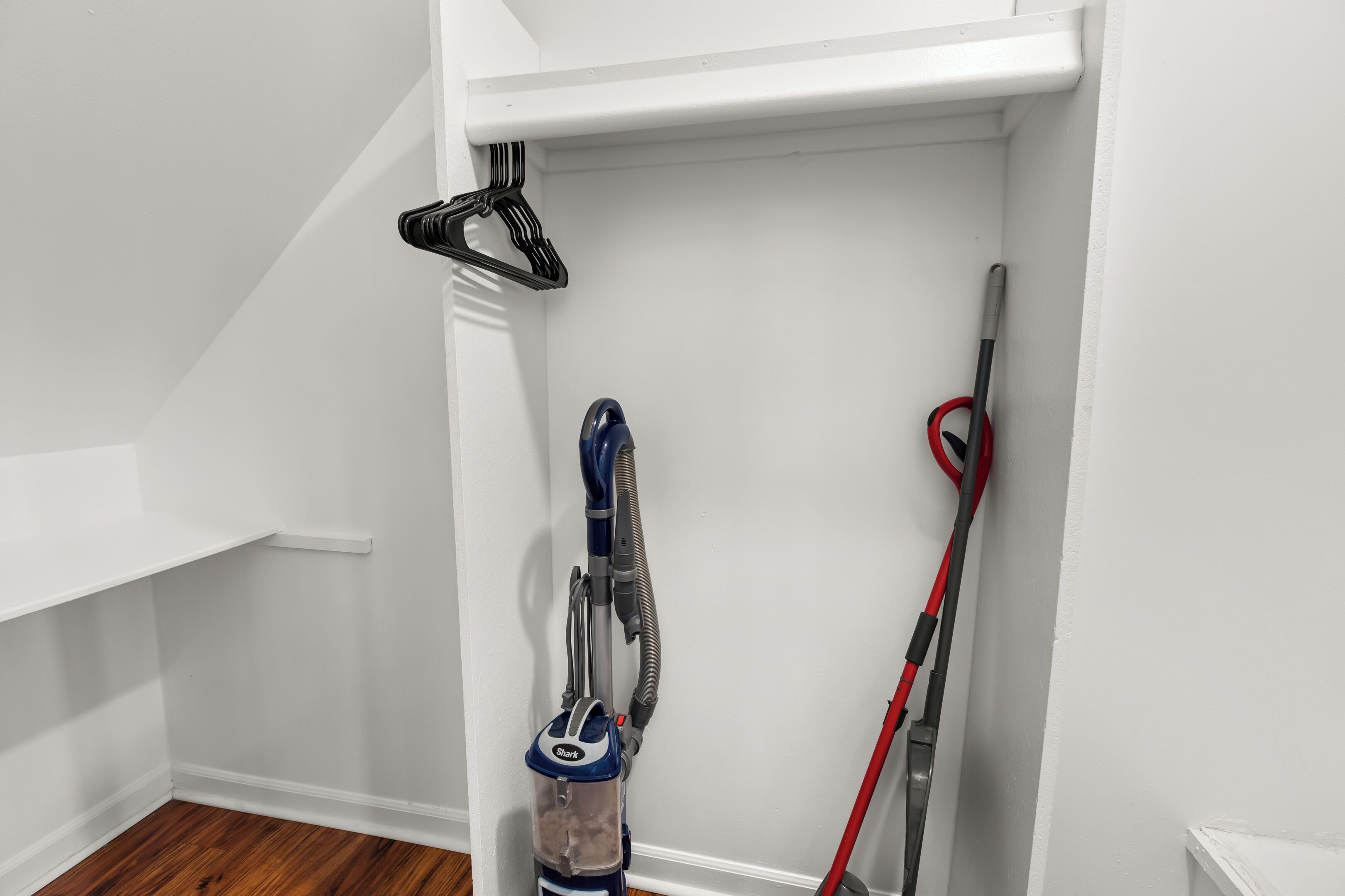 Esquina de una habitación con pared blanca, suelo de madera, una aspiradora y una escoba apoyada en la pared.