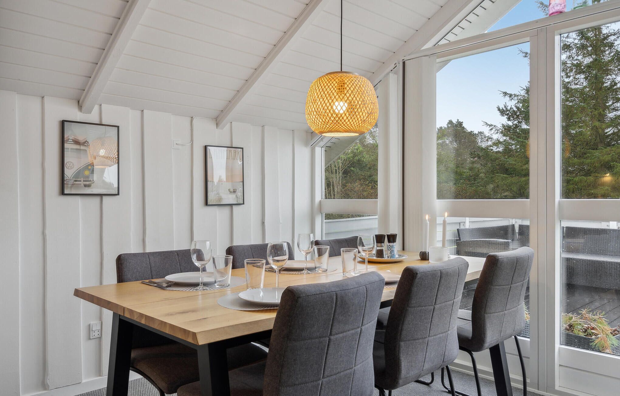 Un espace repas comprenant une table en bois, des chaises à revêtement gris et un luminaire suspendu en fibres tressées.