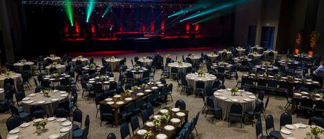 Un salón de banquetes con mesas redondas dispuestas para comer, un escenario con iluminación y suelo alfombrado.