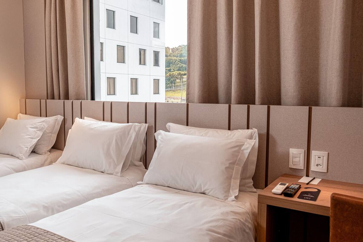Habitación de hotel con dos camas individuales, una mesita de noche de madera con teléfono y control remoto, y una ventana con cortinas.
