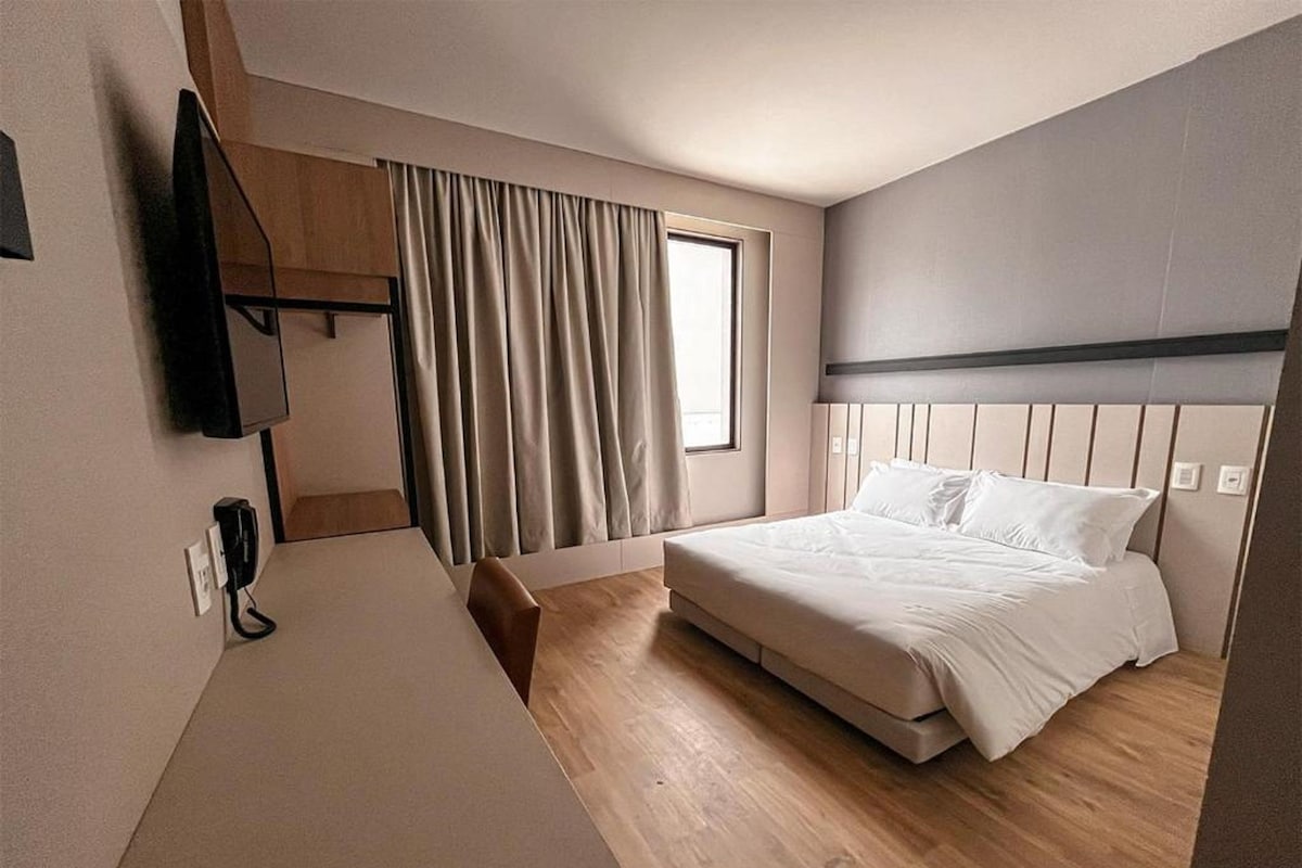 Ein Hotelzimmer mit einem Bett, einem Schreibtisch, einem Stuhl, einem Fernseher und einem fenster mit Vorhängen.