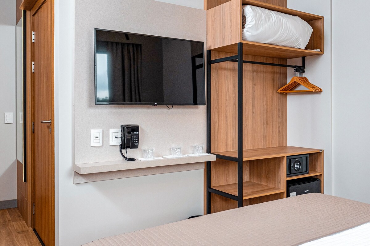 Ein Hotelzimmer mit einem Bett, einem an der Wand befestigten Fernseher, einem Nachttisch mit Telefon und einem Kleiderhaken.