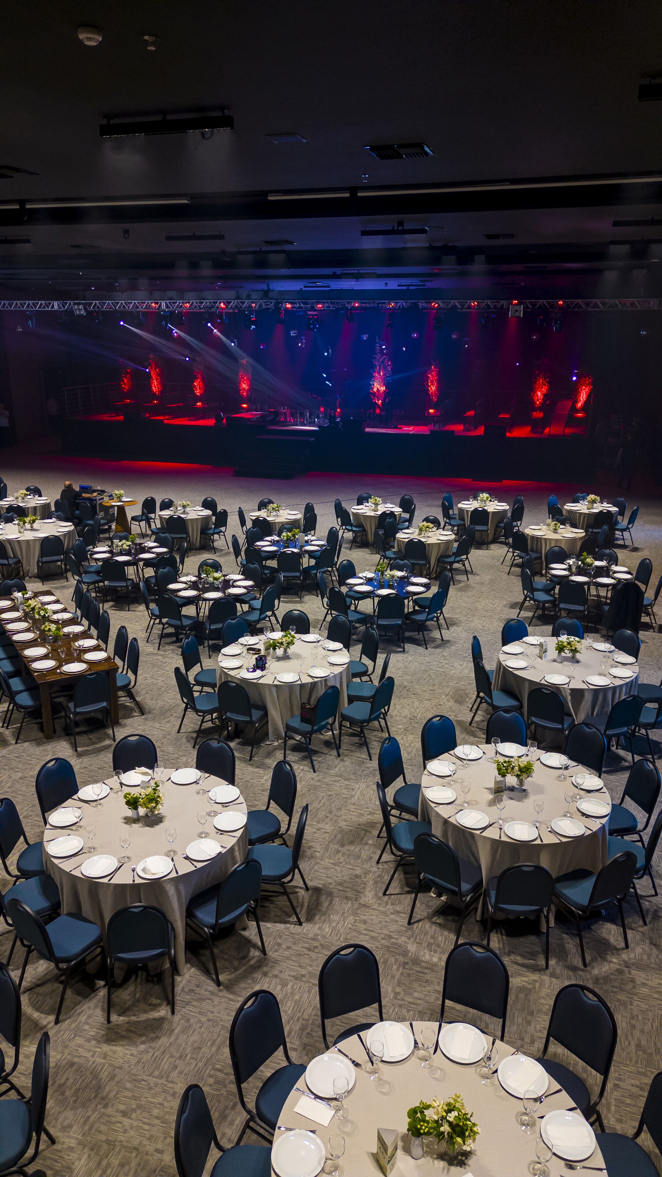 Un salón de banquetes con mesas redondas dispuestas para cenar, un escenario con iluminación roja y suelo alfombrado.