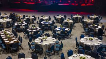 A banquet hall with round tables set for dining, a stage with red lighting, and a carpeted floor.