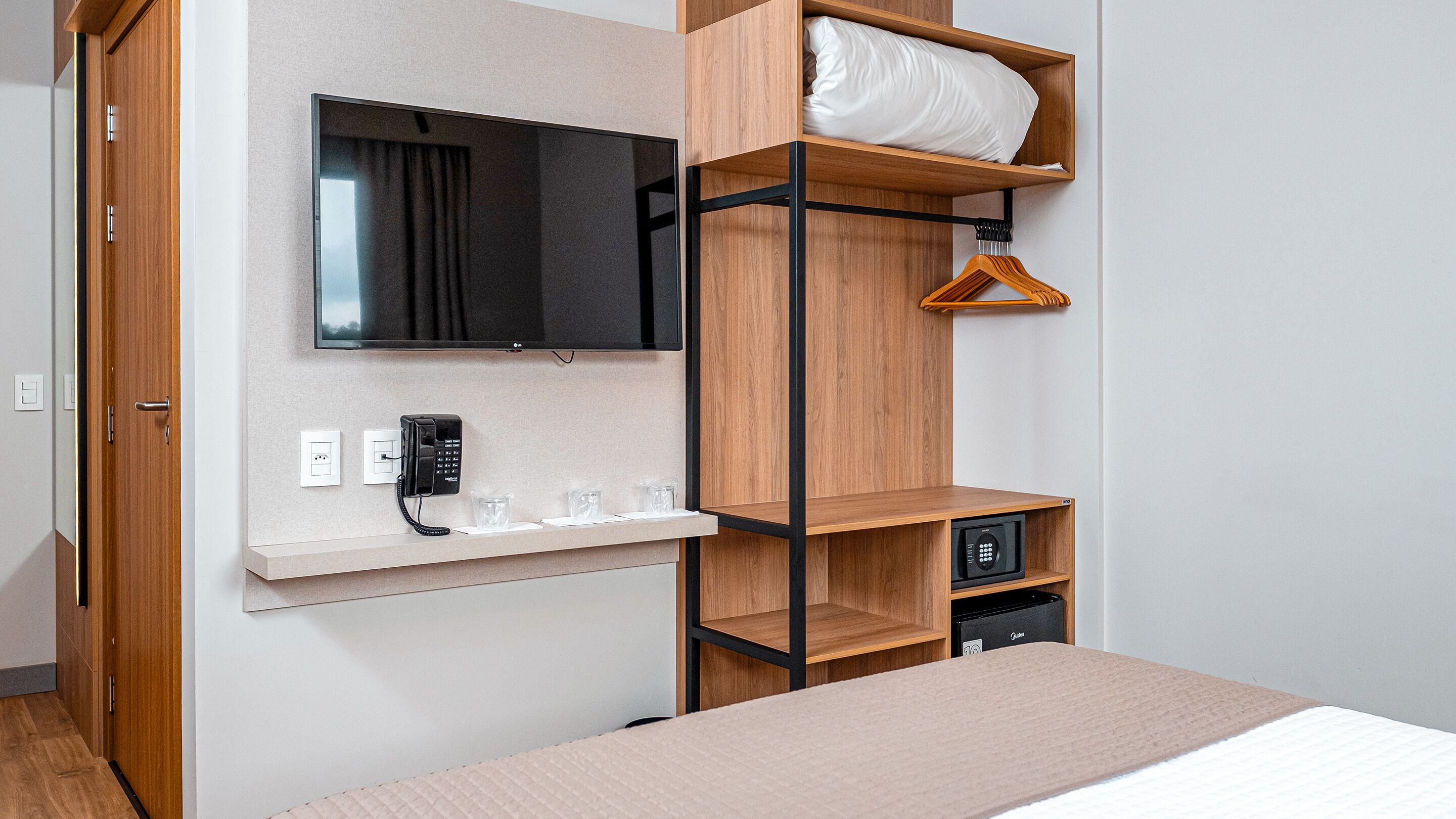 A hotel room with a bunk bed, a flat-screen TV mounted on the wall, a bedside table with a phone and glasses, and a small safe.