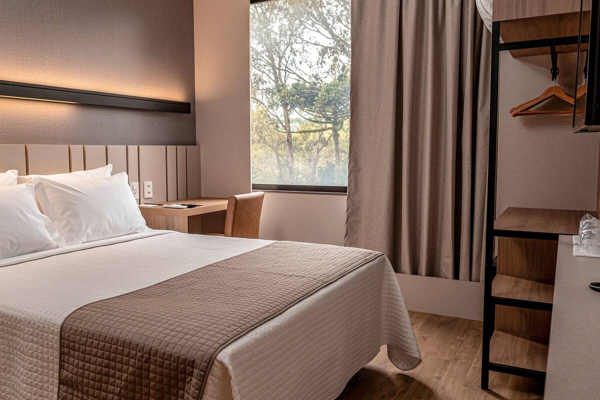 Habitación de hotel ordenada con cama, mesita de noche, silla y escritorio con perchero.