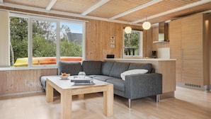 A modern living room with a wooden ceiling, a gray sofa, a wooden coffee table, and a view of greenery outside.