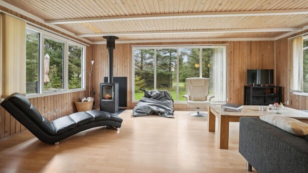 A cozy living room with a curved black leather sofa, a grey armchair, a wooden coffee table, and a TV stand. A black wood-burning stove is centrally located, and large windows offer a view of the greenery outside.
