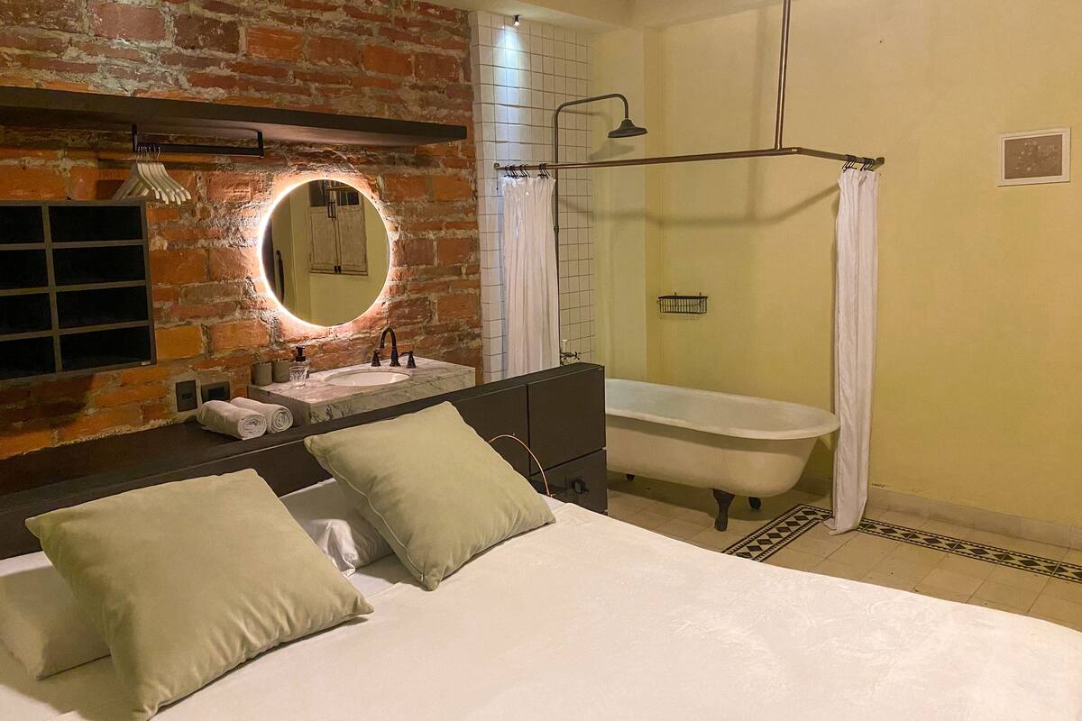 A bedroom with a bed, two pillows, a nightstand, a mirror, a vanity with a sink, and a bathtub.