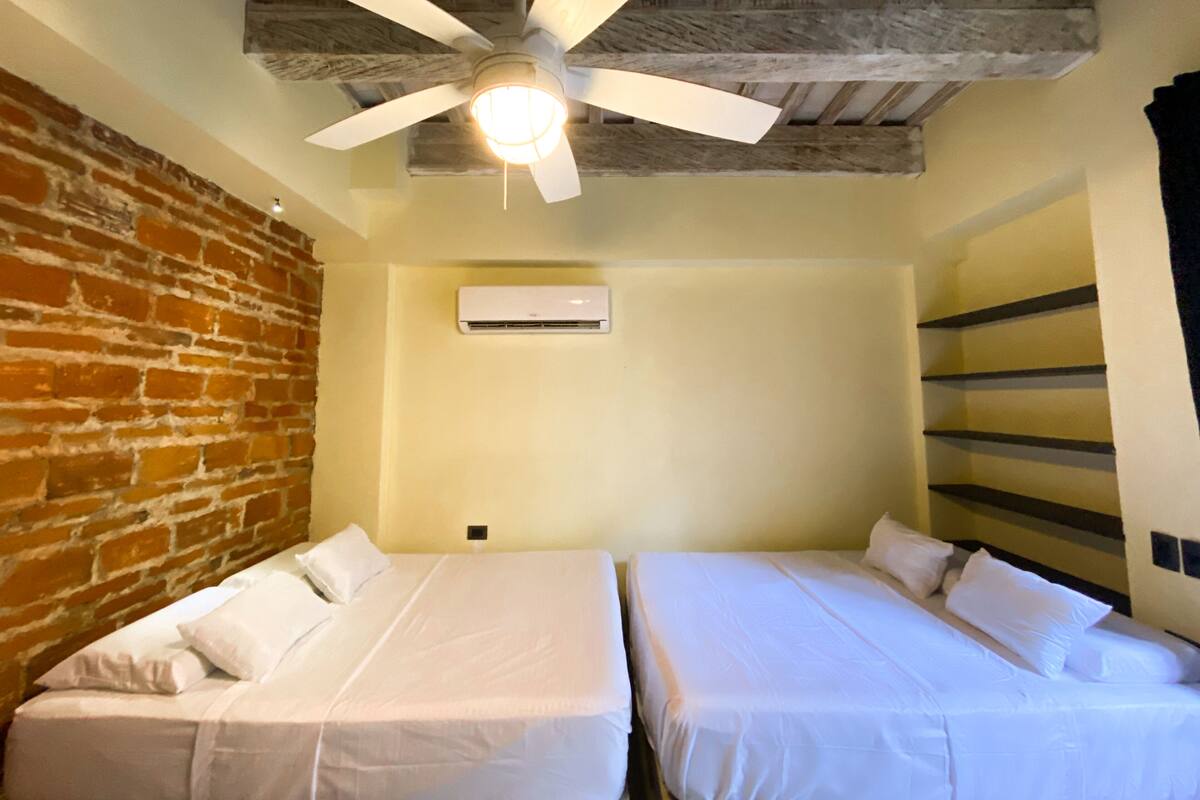 A room with two single beds, a ceiling fan, an air conditioning unit, and exposed wooden beams.