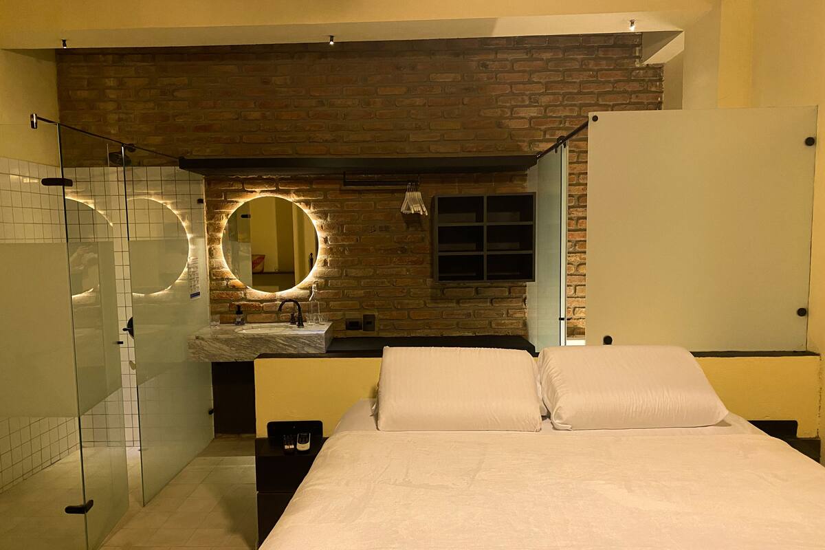 A hotel room with a brick wall, a large bed, a bathroom with a mirror, and a shower.