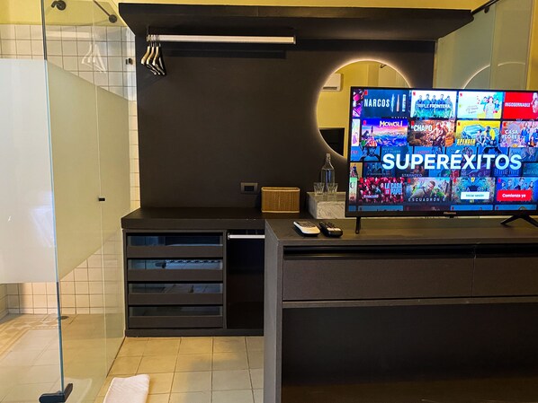Un baño moderno con un espejo grande, un tocador con lavabo y un televisor mostrando diferentes programas.