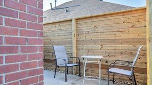Un patio al aire libre con dos sillas de metal y una mesita blanca, rodeado por una cerca de madera y un muro de ladrillo.