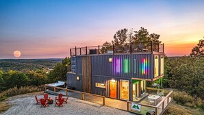 A modern, multi-level cabin with a deck, outdoor seating, and a scenic view of the landscape.