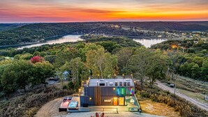 A modern cabin with a deck, outdoor seating, and a view of a lake and hills at sunset.