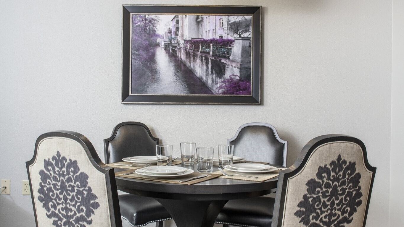 Ein Esstisch ist gedeckt mit Tellern, Gläsern und Besteck, begleitet von gepolsterten Stühlen mit dekorativen Rückenlehnen, vor einer Wand mit einem gerahmten Bild.