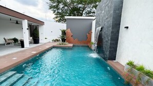 Una piscina con una cascata, circondata da un muro bianco e un solarium in mattoni.